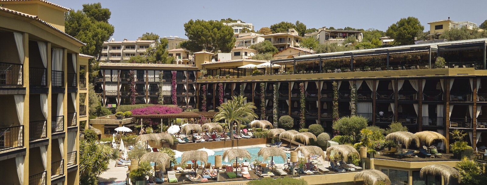 Aerial drone view of Bikini Island & Mountain's tiered hotel wings around a central pool with thatched-roof cabanas, hillside and mountains behind