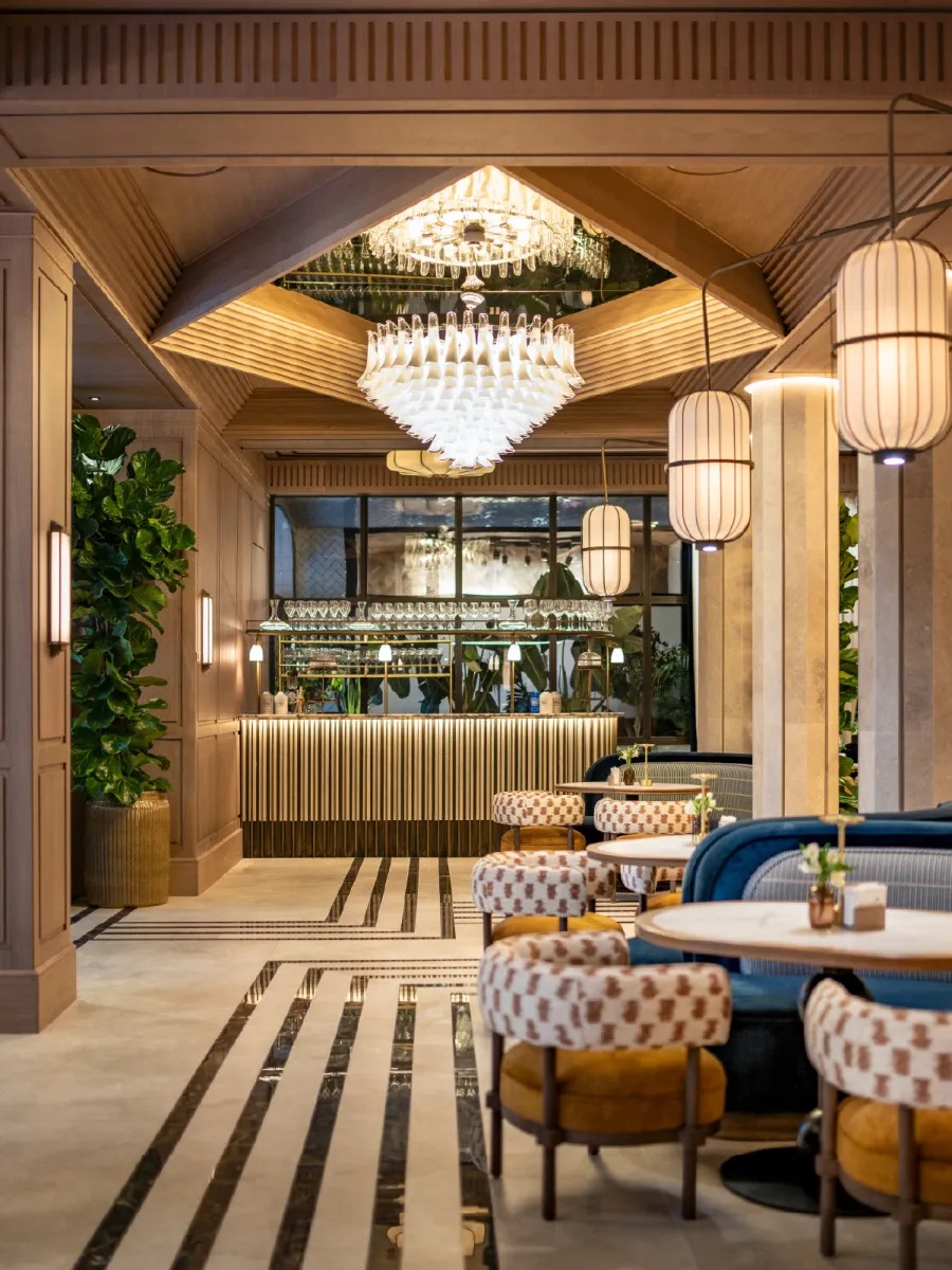 The bar with a fluted, under-lit counter, shelves of glassware against a mirrored back wall, a glass-petal chandelier and patterned stools.