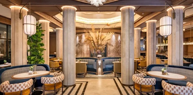 A restaurant-lounge interior with stone columns, blue velvet banquettes, patterned armchairs and white paper lanterns around marble-topped tables.