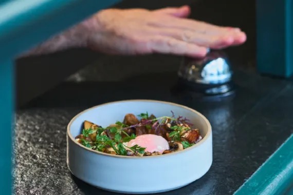 A ceramic bowl of sauteed mushrooms and herbs topped with a soft egg, set on a dark stone counter beside a service bell.