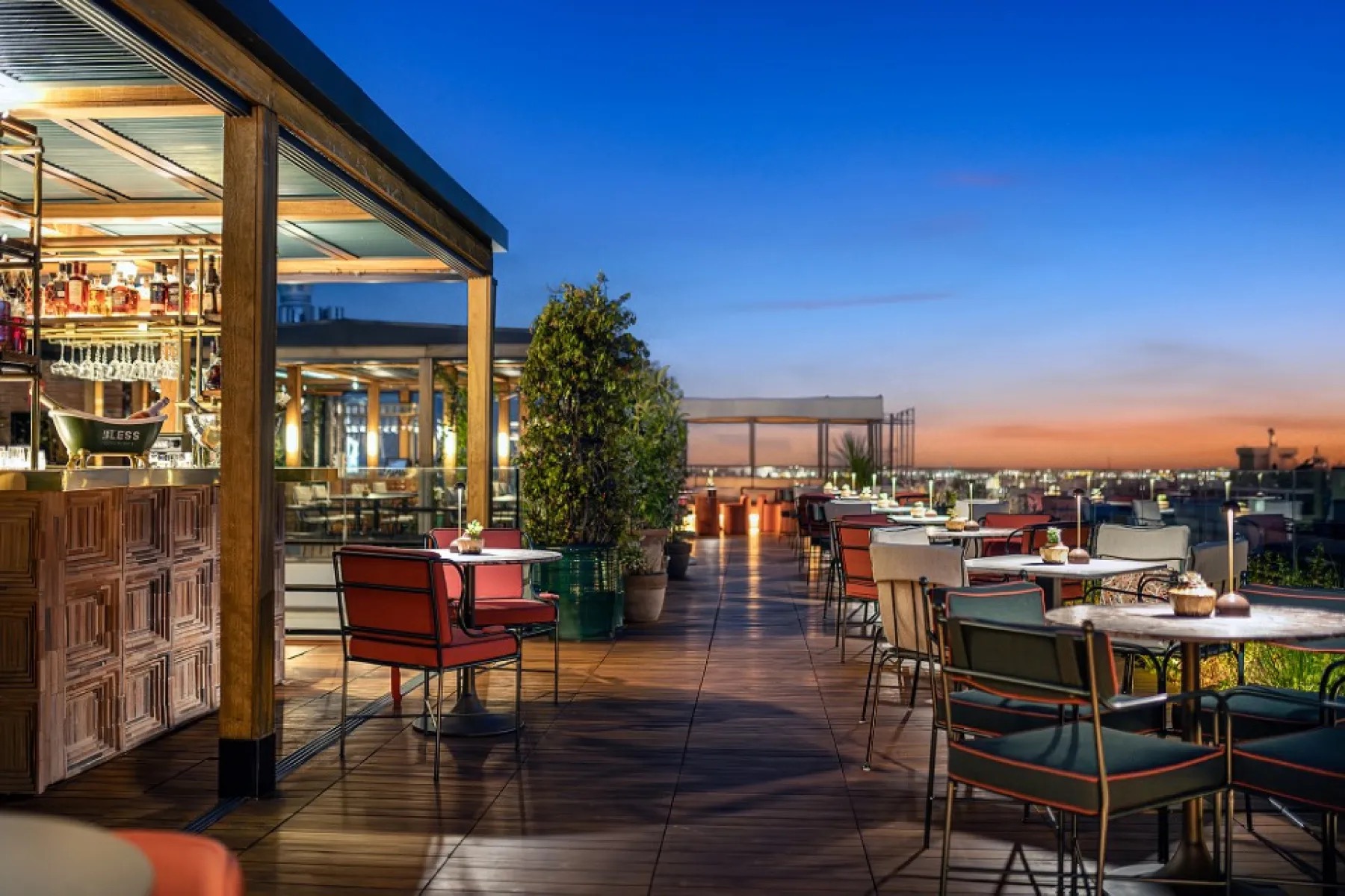 The rooftop terrace at dusk: a wooden deck with marble bistro tables, red and green chairs, potted trees and a corner bar with a BLESS champagne bucket.