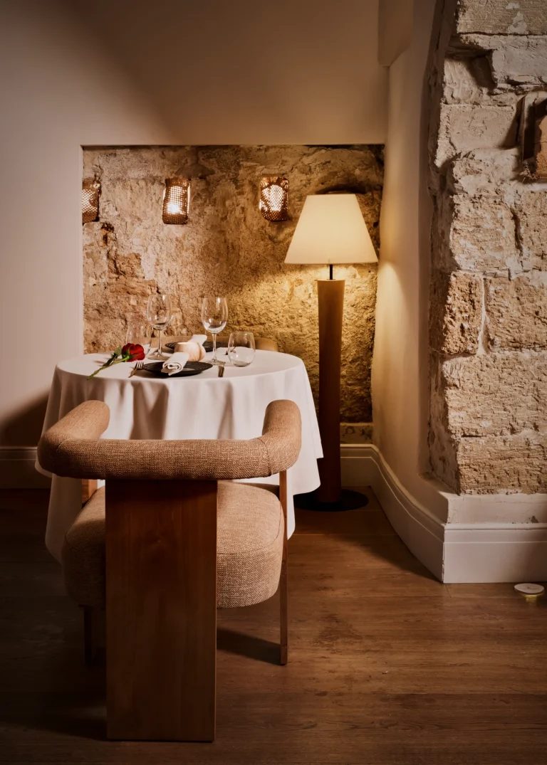 Candlelit two-top table for dinner at Posada Terra Santa's restaurant against an exposed stone wall, set with wine glasses and a single rose