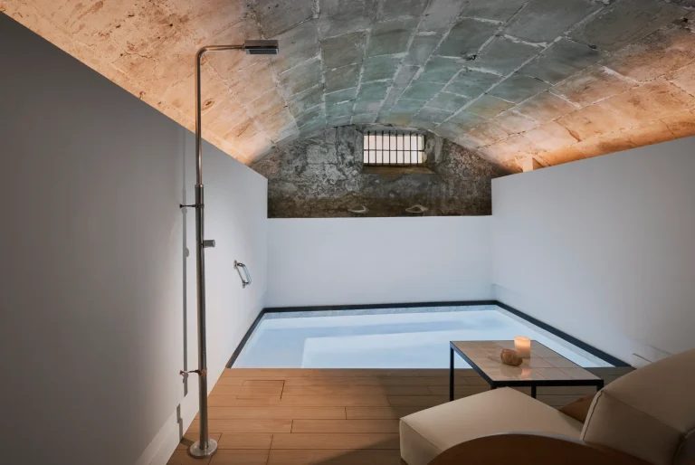 Small plunge pool at Posada Terra Santa set under a stone vaulted ceiling, with a freestanding shower, wood decking and a low table