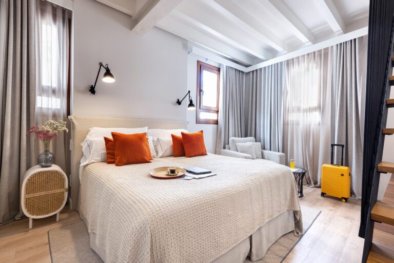 Guest room at Posada Terra Santa with a white beamed ceiling, orange throw pillows on the bed, a rattan side table and a breakfast tray in bed
