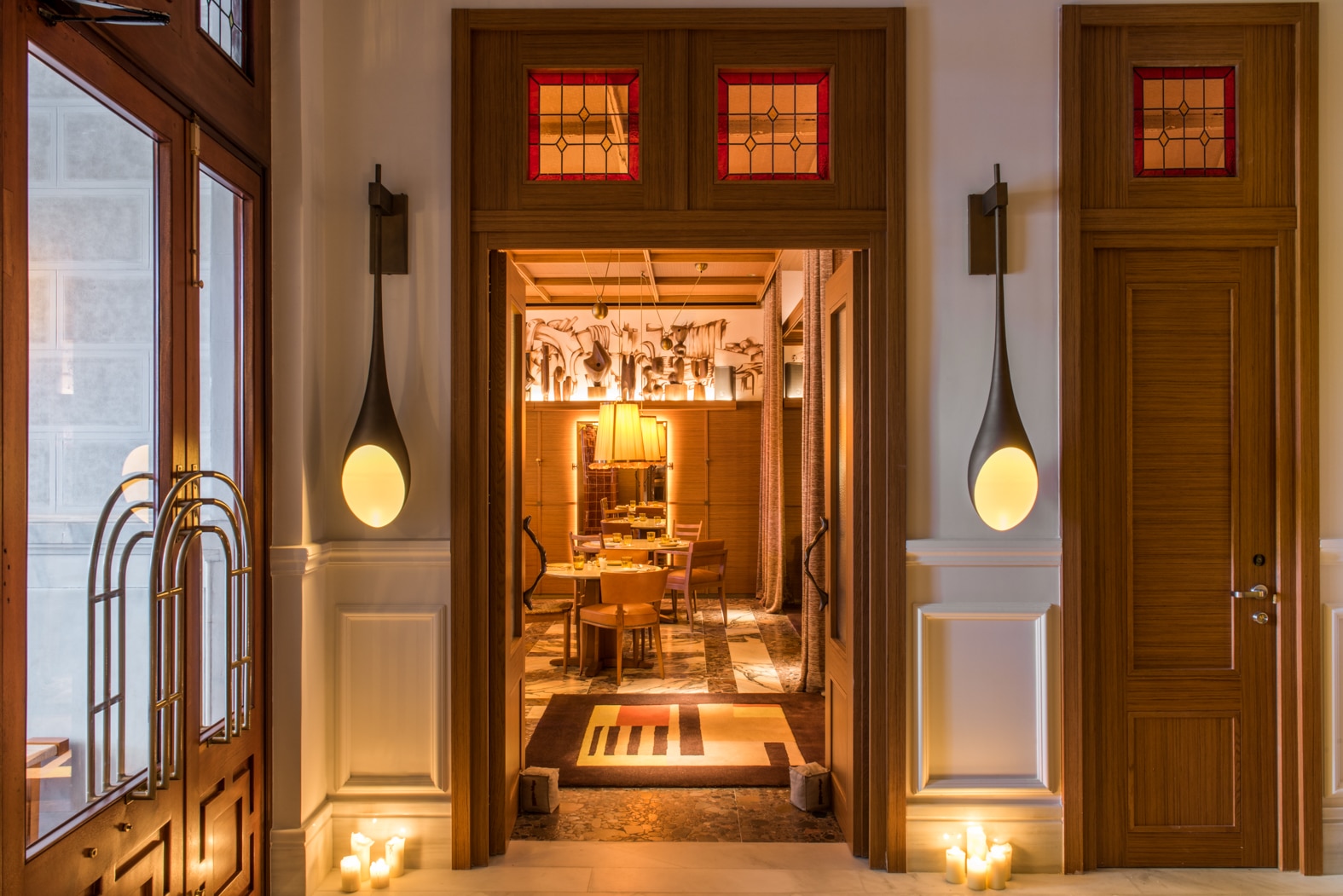 The restaurant entrance: wooden double doors with red stained-glass transoms open onto a dining room with round tables, wooden chairs, a marble floor, a pleated pendant lamp and a frieze of sketched figures, with teardrop wall lights and candles either side.