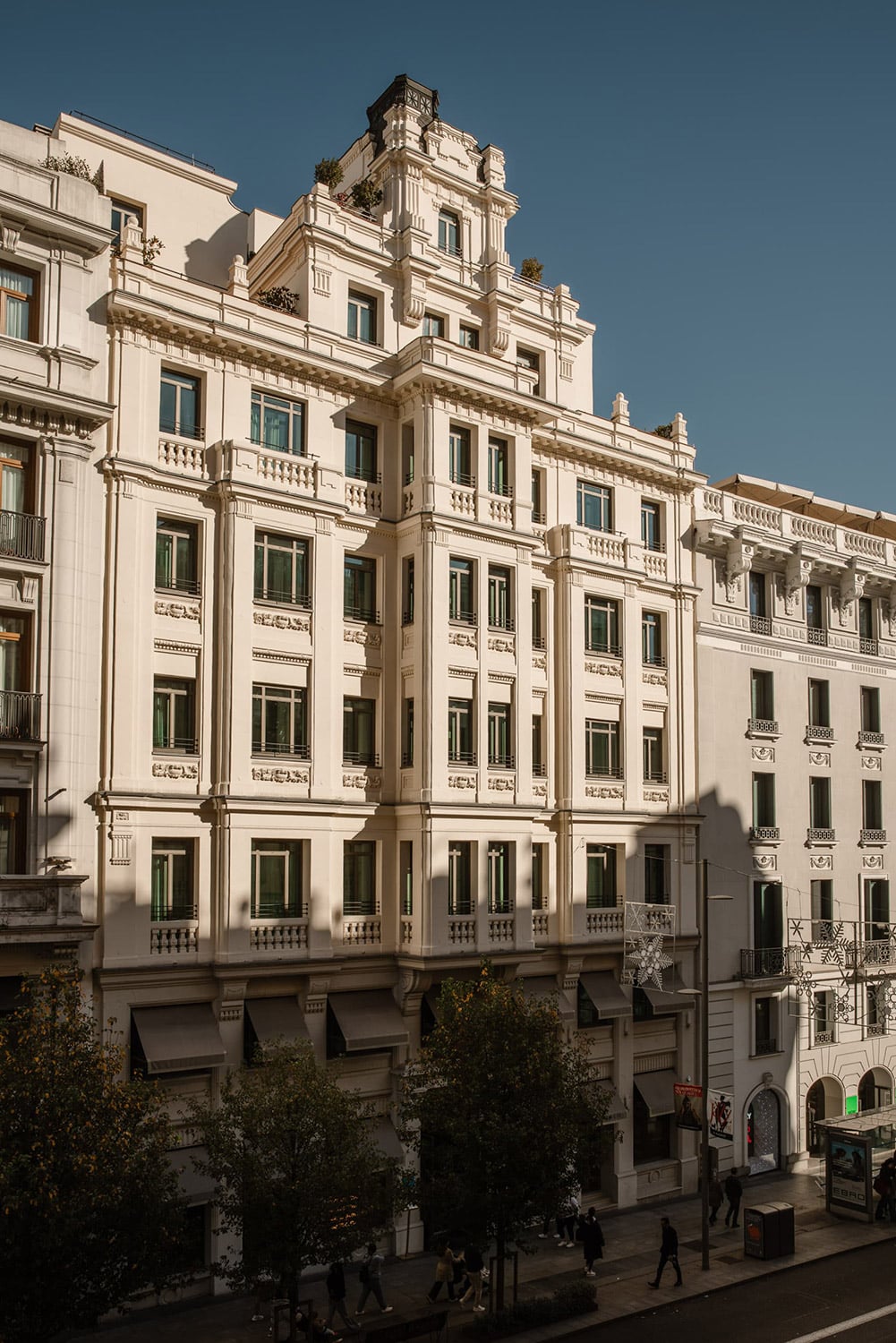 The hotel's white, ornately moulded facade seen from across the street: rows of balconied windows, planted terraces at the top, black awnings over the ground floor and star-shaped lights strung across the road.