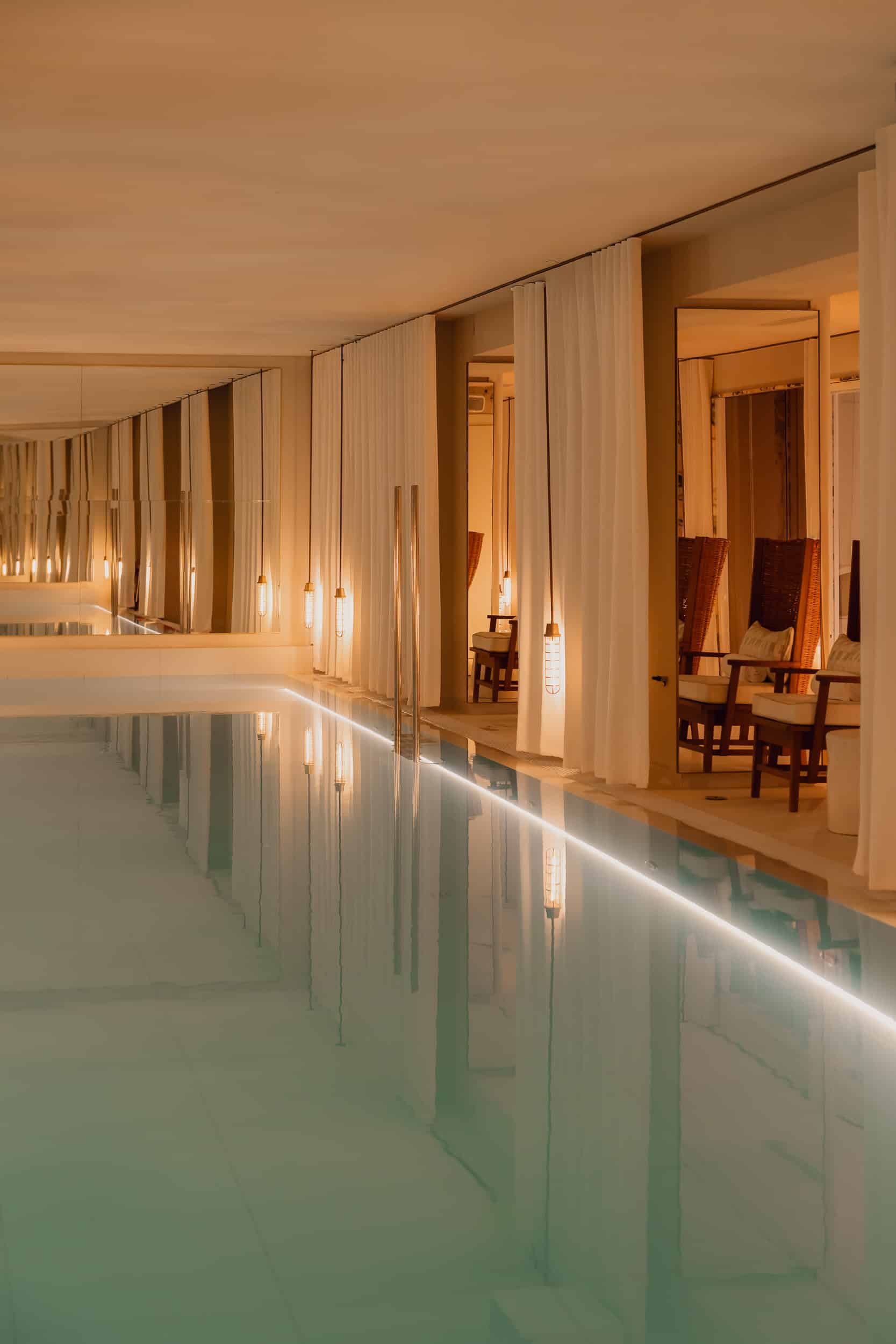 The indoor pool: a long pale-green lane lit by an LED strip along its edge, white curtains between mirrored piers and rattan-backed loungers with white cushions in alcoves beside it.