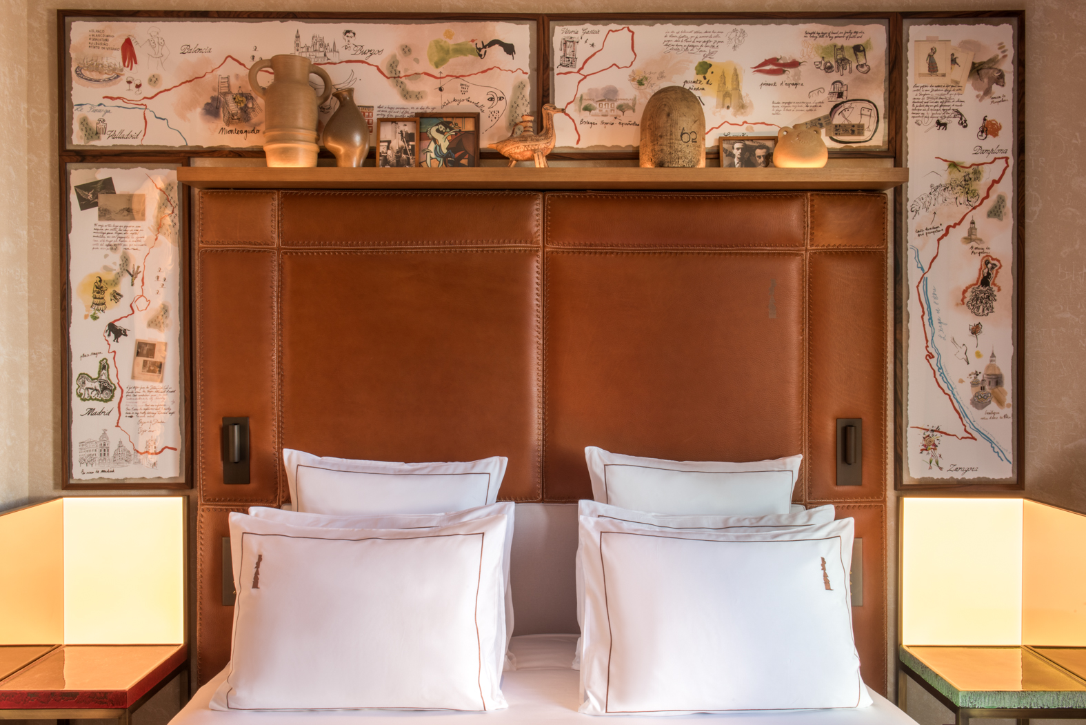 A guest room bed with a stitched tan leather headboard and four white pillows, flanked by illuminated bedside cubes and framed illustrated maps of Spanish cities including Madrid, with a shelf of ceramics and small pictures above.