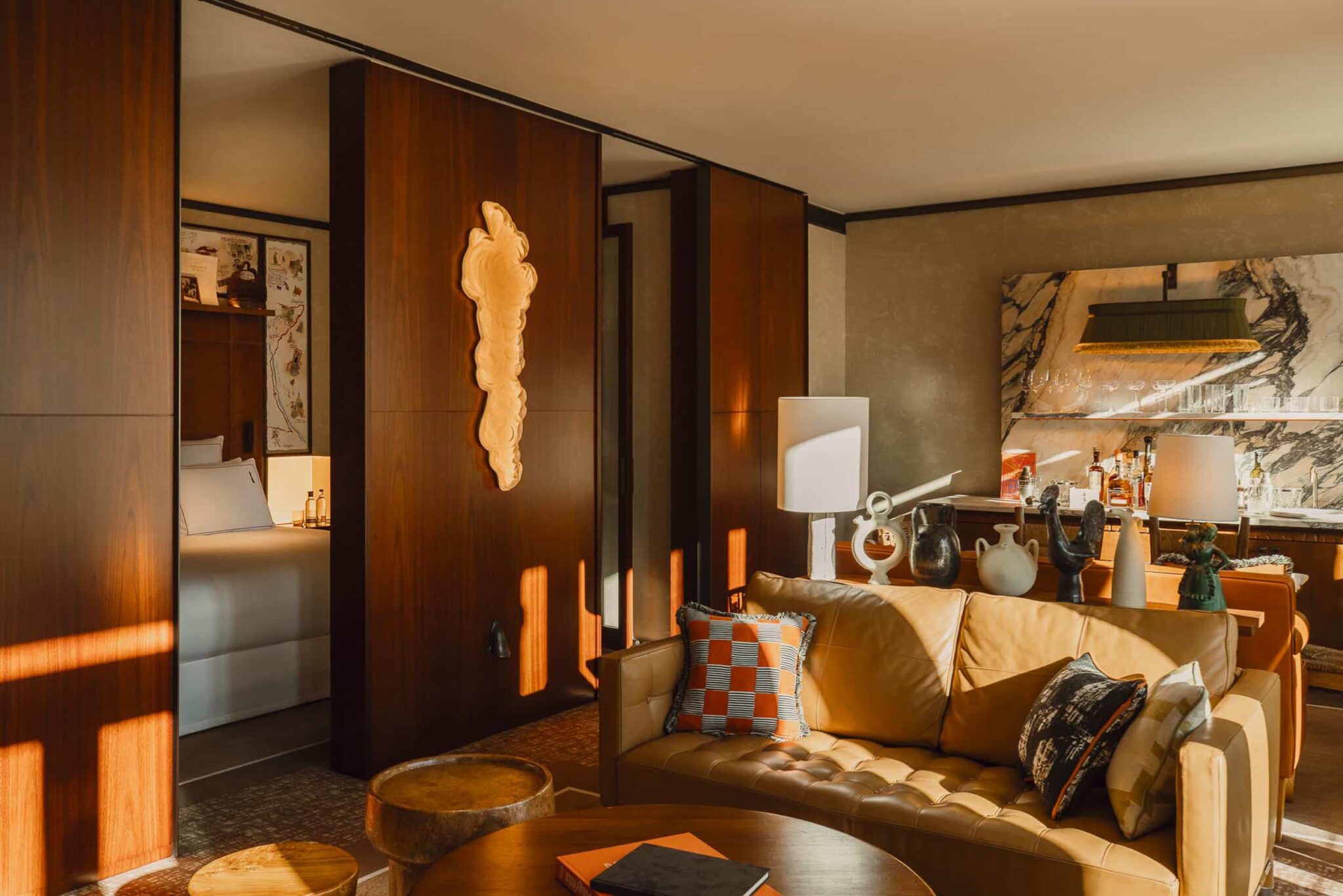 A sunlit suite living room: a tan tufted leather sofa with patterned cushions, walnut sliding panels open to a bedroom with a leather headboard, a bar cabinet with glassware against veined marble, and ceramic vessels on a shelf.