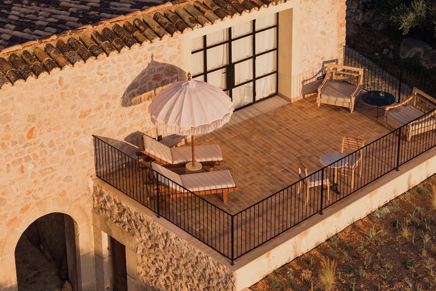 A raised timber terrace on the stone building, with sun loungers, a fringed parasol and cane armchairs