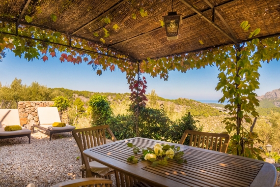 Vine-covered pergola terrace with lounge seating and a dining table, overlooking the hills toward the coast
