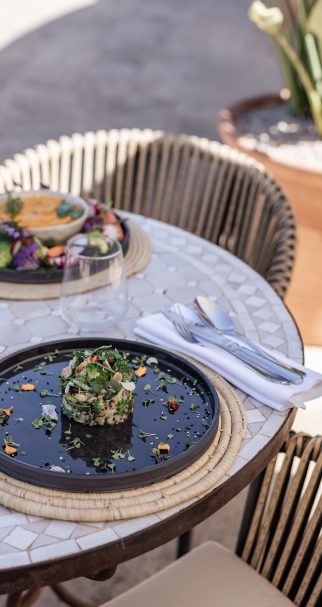 Poolside restaurant table at Cal Reiet with a mosaic tabletop, rattan chairs and a plated dish, pool edge visible behind