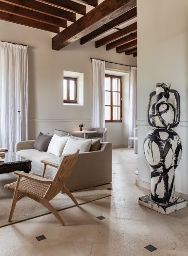 Entrance lounge at Cal Reiet with exposed dark wood beams, a cream sofa, a rattan armchair and a black-and-white abstract sculpture on the stone floor