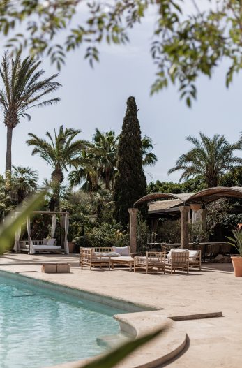 Outdoor pool at Cal Reiet framed by palm and cypress trees, with a stone patio and a pergola with lounge seating beyond
