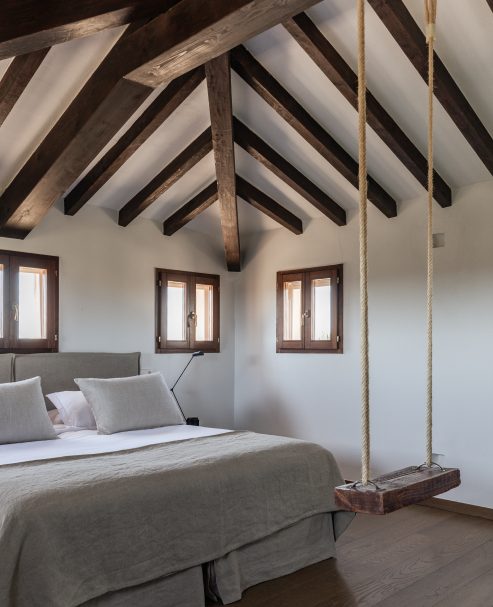 Attic guest room at Cal Reiet with exposed dark wood A-frame beams, grey linen bedding and a rope swing hanging in the corner