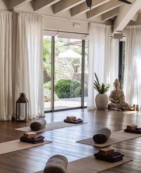 Indoor yoga shala at Cal Reiet with a wood beam ceiling, sheer white curtains, mats and bolsters laid out and a Buddha statue in the corner