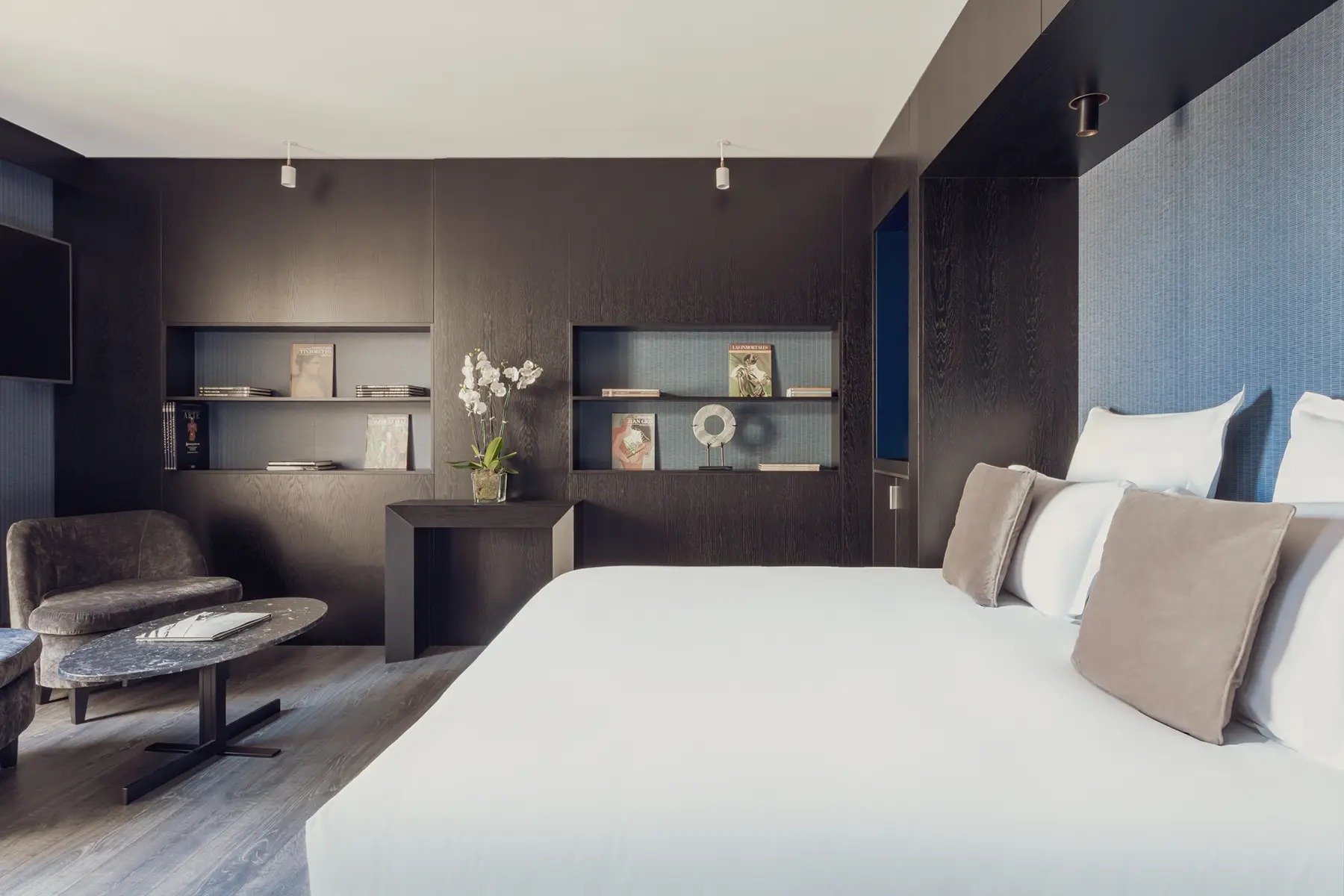 King bed with white linen in a room with dark wood shelving, a blue padded headboard wall, an armchair and an oval stone coffee table