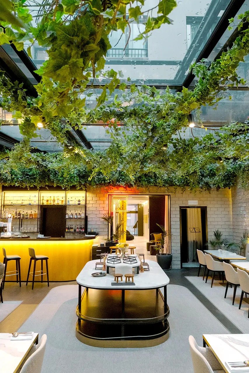 Glass-roofed interior garden dining area under hanging ivy, with a lit yellow bar, a marble-topped table and set tables