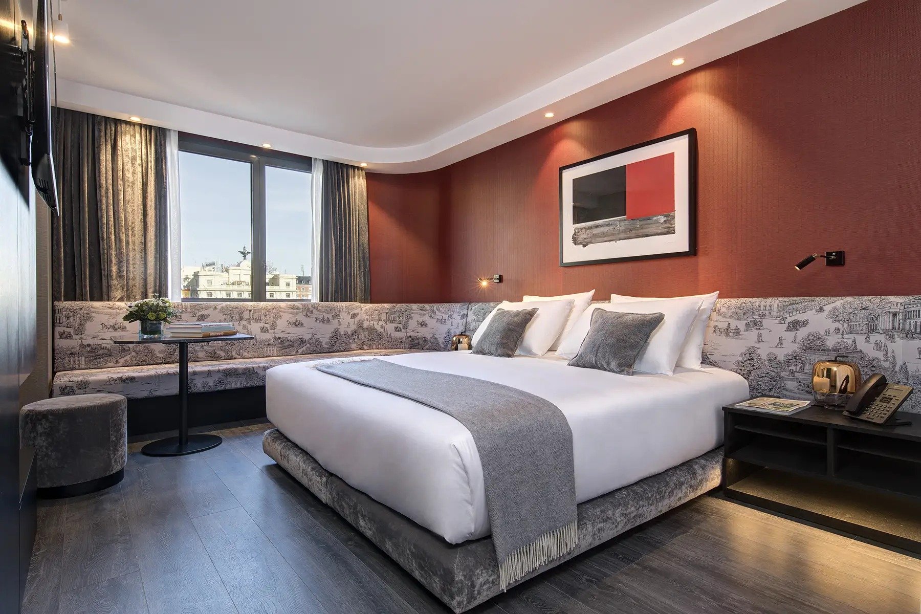 King bed with a grey throw in a room with a red feature wall, a framed abstract print, toile-patterned banquette seating and a window
