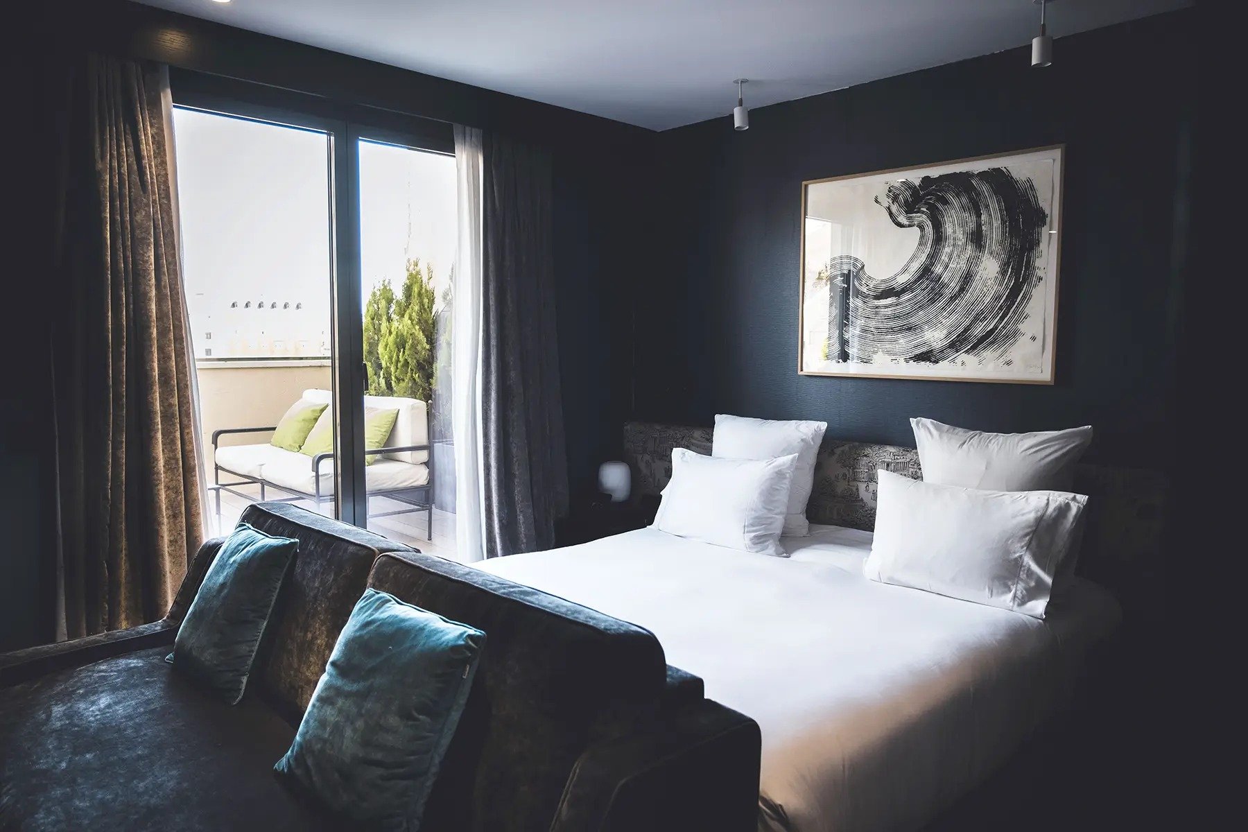 Dark-walled bedroom with a king bed, velvet sofa and a framed black-and-white print, with a sliding door onto a terrace with outdoor seating