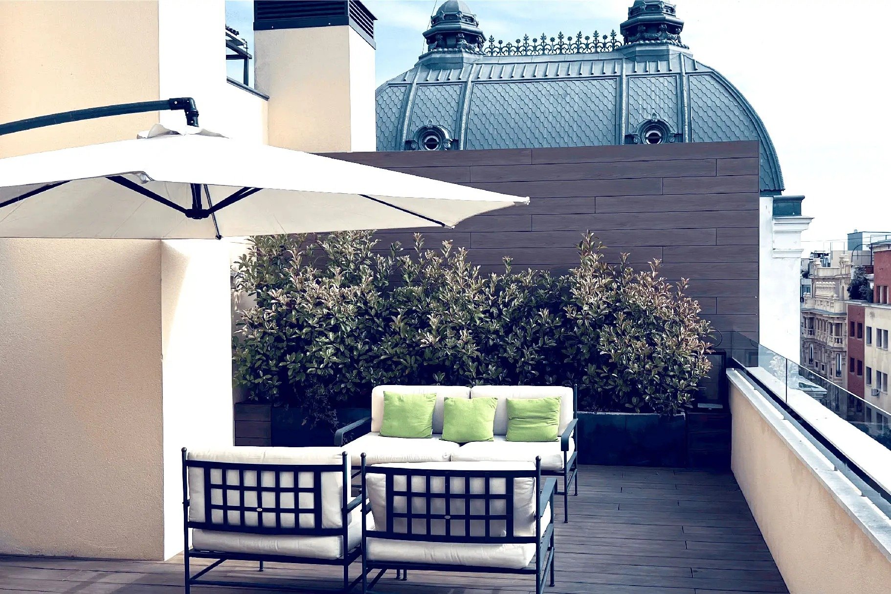 Rooftop terrace with white cushioned outdoor seating, a cantilever parasol and a planted hedge, with a slate-roofed dome behind
