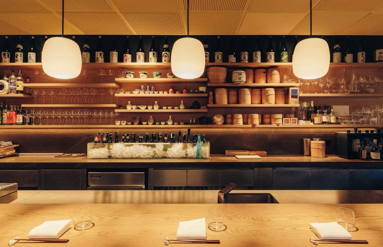 Wooden counter and shelves lined with sake bottles and glassware under pendant lamps at the Dos Palillos bar inside Casa Camper Barcelona.