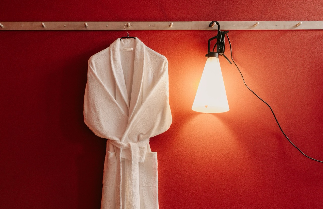 White bathrobe on a hook and a lit cone-shaped lamp against a red wall in a room at Casa Camper Barcelona.