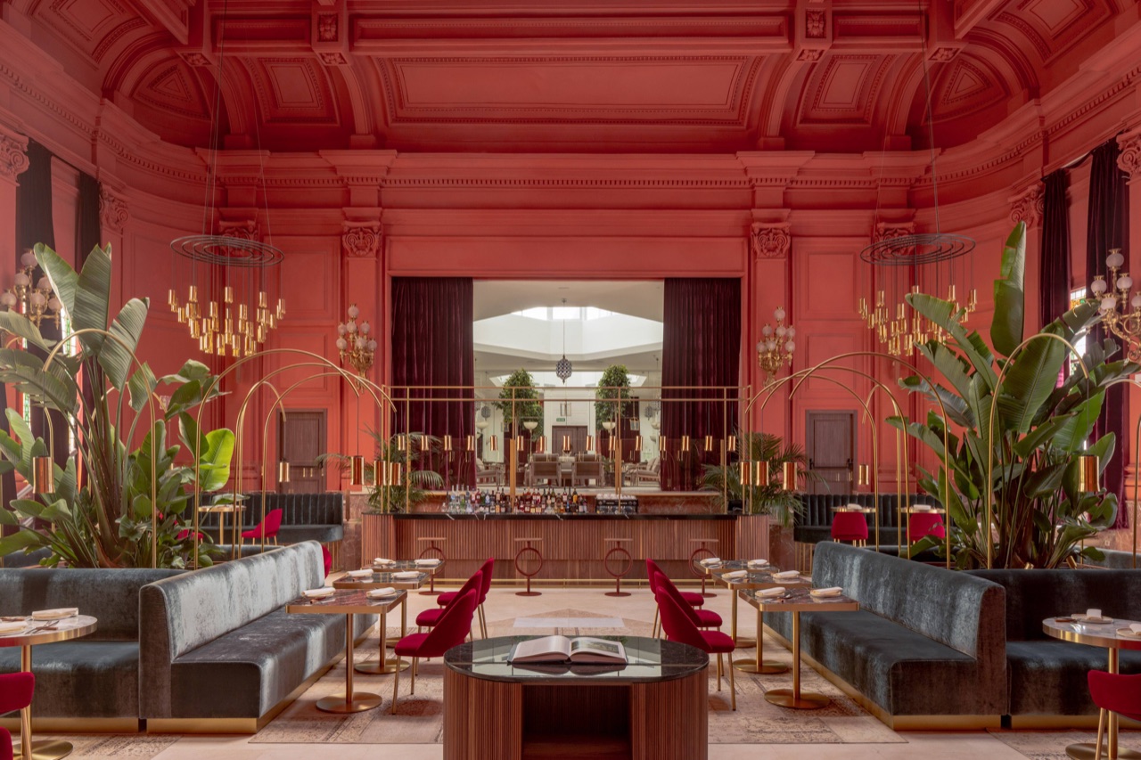 Coral-red domed dining hall with a central bar, velvet banquettes, palm plants and brass chandeliers, part of the Maché restaurant.