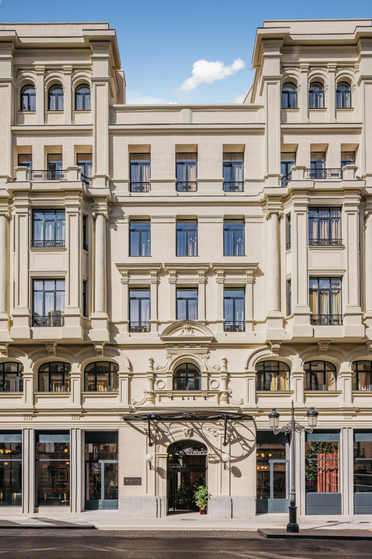 Cream stone hotel facade with ornate balconies and arched windows across several floors, street-level entrance under a metal and glass canopy.