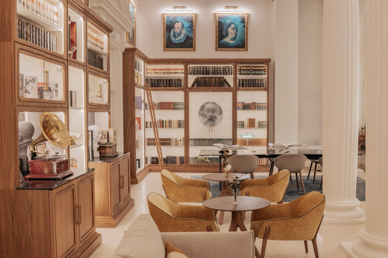 Two-storey library lounge with built-in wooden bookshelves, a rolling ladder, mustard-yellow armchairs and round tables.