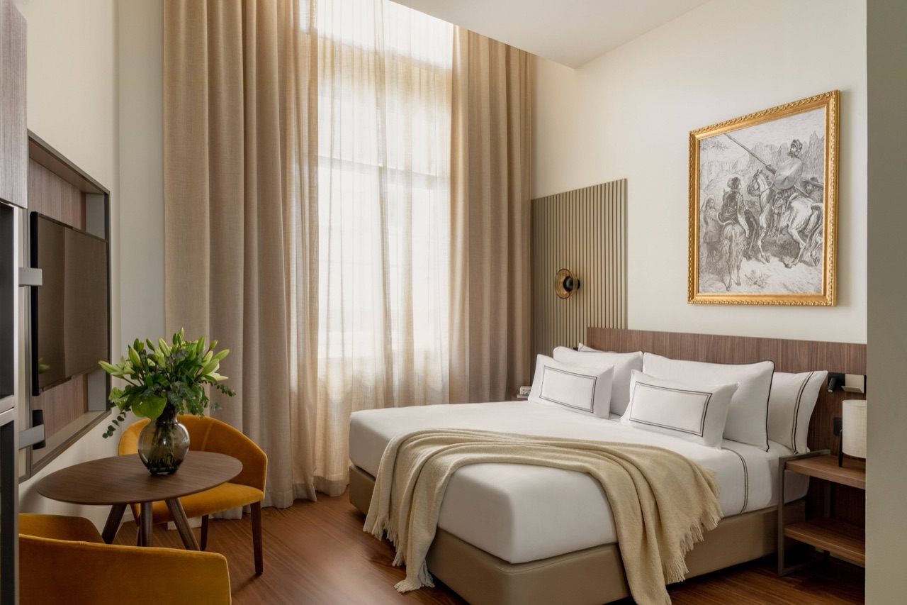 Hotel bedroom with a white made bed, beige throw, tall curtained window and a framed black-and-white illustration of Don Quixote on horseback.