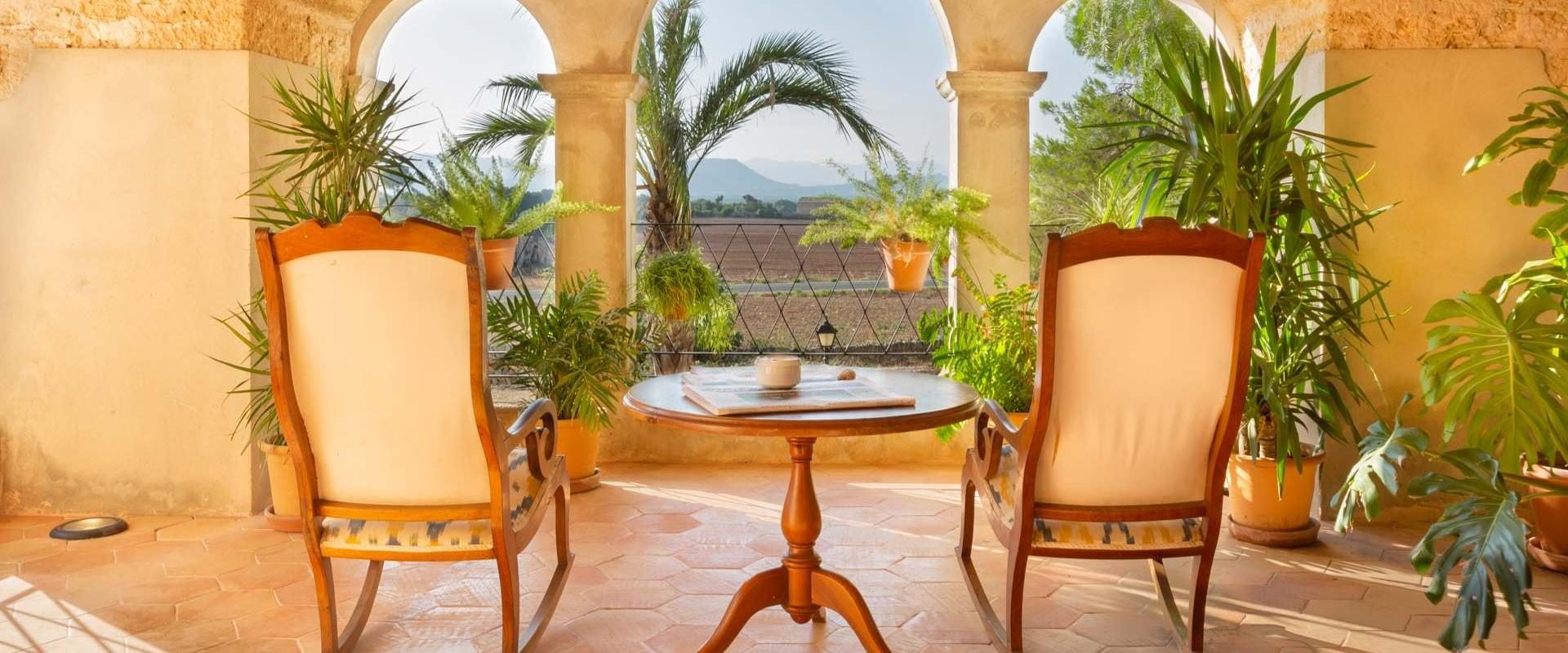Arched stone loggia at Casal Santa Eulalia with wicker armchairs around a round wood table, potted palms, and a view over Mallorcan farmland