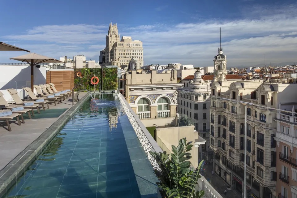 A rooftop infinity pool lined with sun loungers, overlooking Gran Via rooftops and the Telefonica building in the distance.
