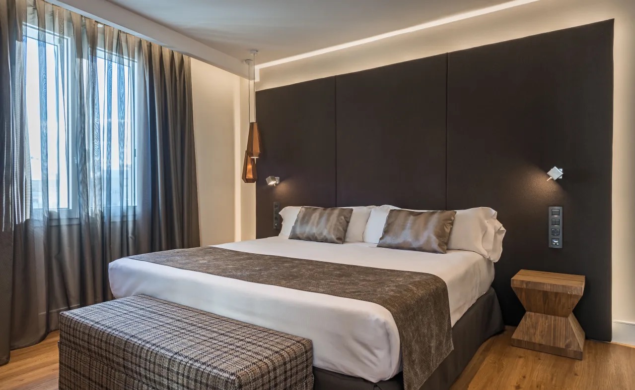 A modern hotel room with a dark brown upholstered headboard, hanging pendant light and a wooden side table.