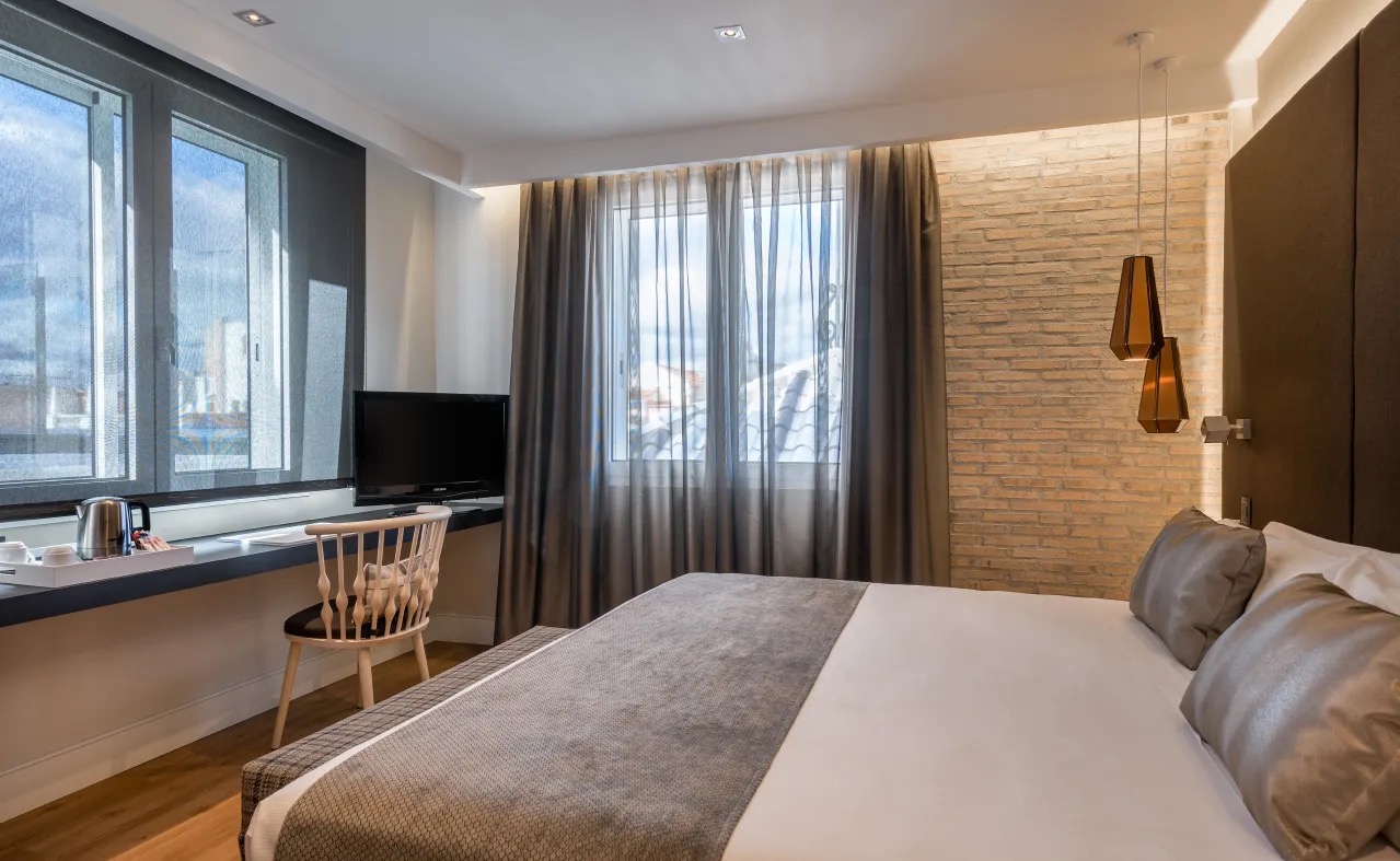 A hotel room with an exposed brick accent wall, a wall-mounted TV, a work desk by the window and a kettle tray.