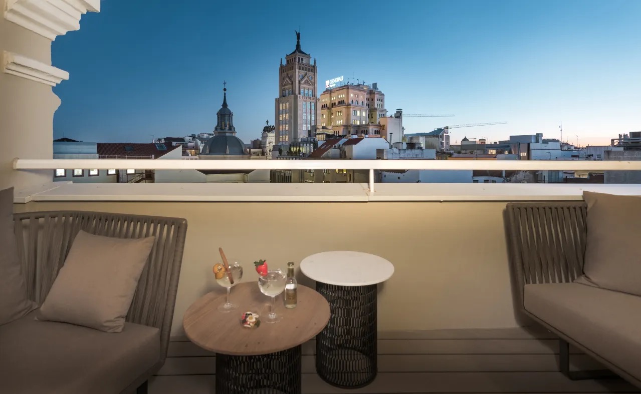 A private room terrace at dusk with two lounge sofas, a round table holding drinks, and Gran Via's skyline including a lit clock tower in the background.