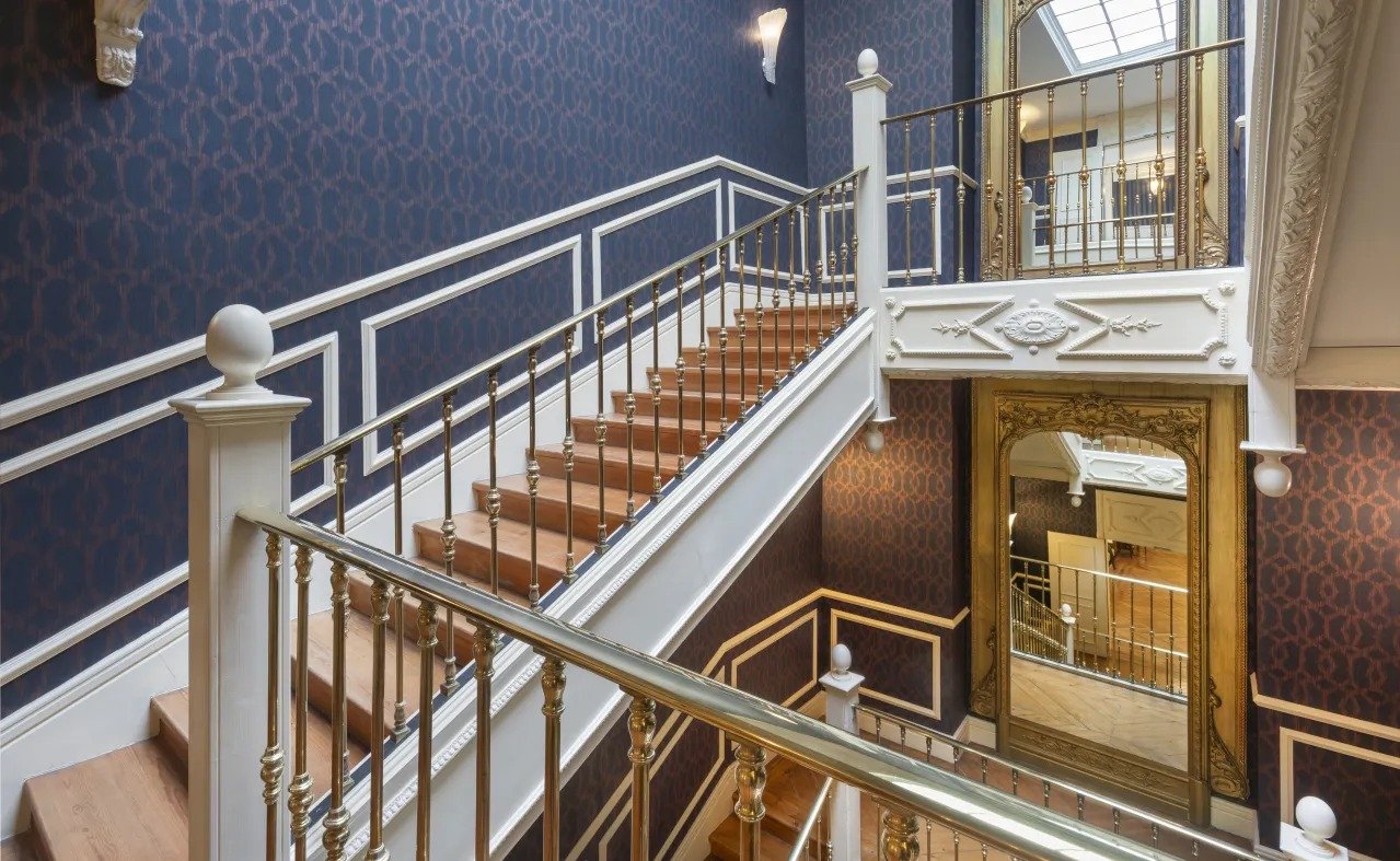 The main staircase at Catalonia Las Cortes, with brass balusters, white moulded panels, navy patterned wallpaper and a gilt-framed mirror.