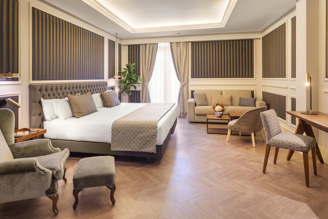 A superior room with a large bed with a tufted headboard, striped wall panels, a sofa, armchair and desk on a herringbone floor.