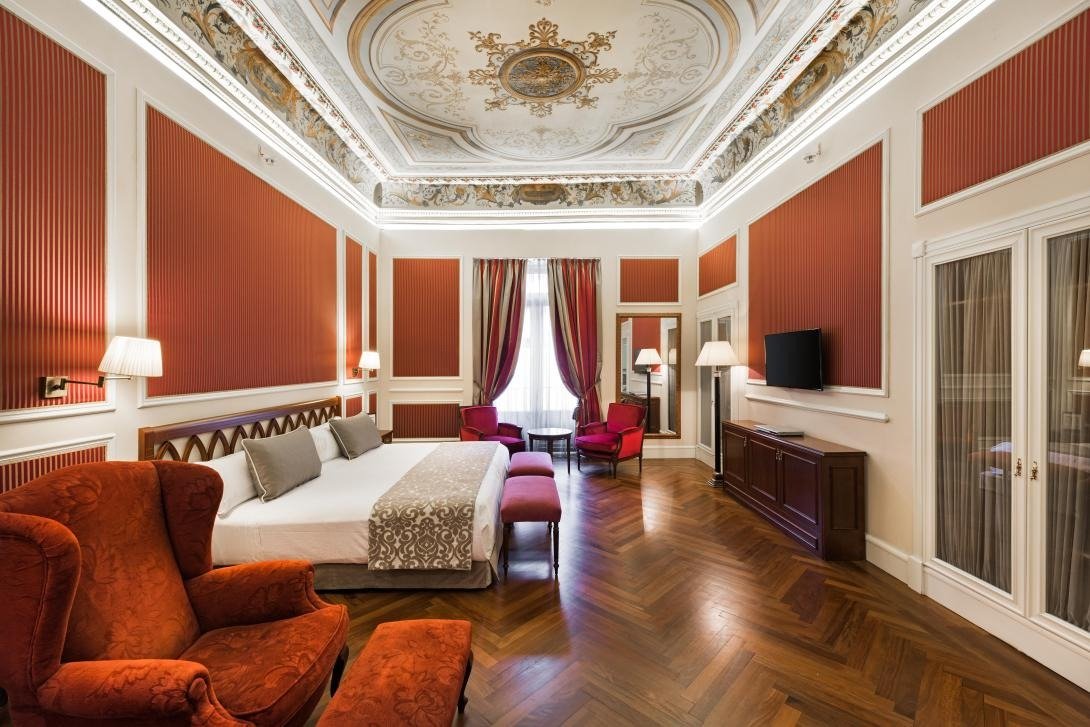 A Catalonia Las Cortes junior suite with a painted, moulded ceiling, red striped wall panels, a double bed, red armchairs and a herringbone parquet floor.