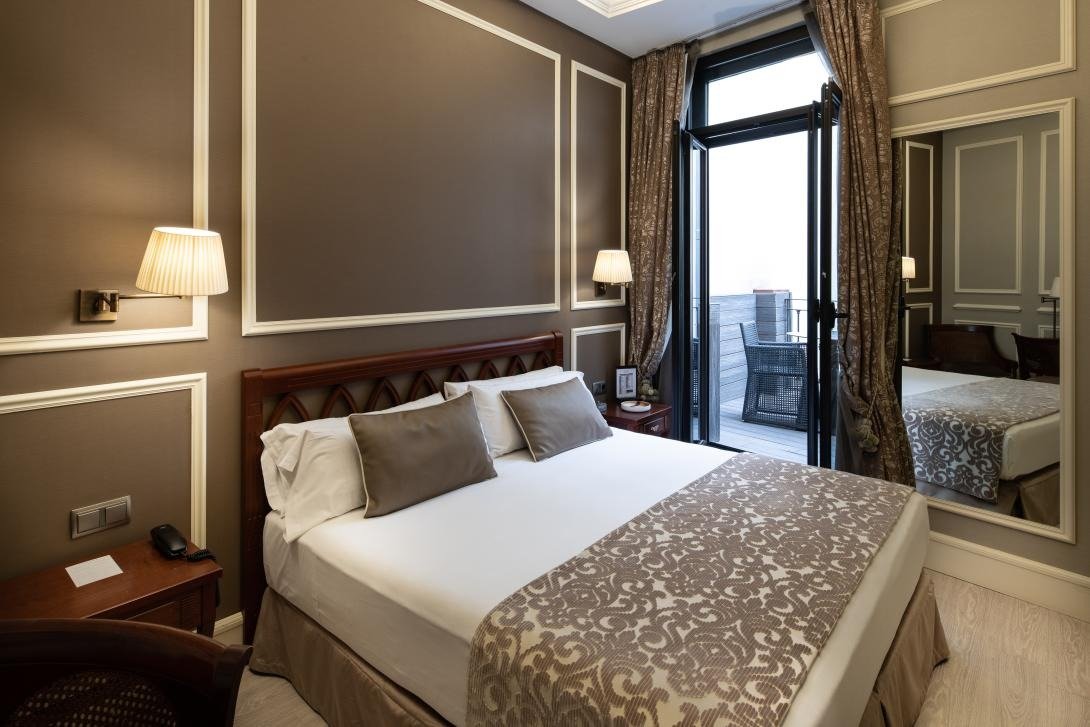 A terrace room with a double bed, brown panelled walls and glass doors opening onto a small wooden terrace with a wicker chair.