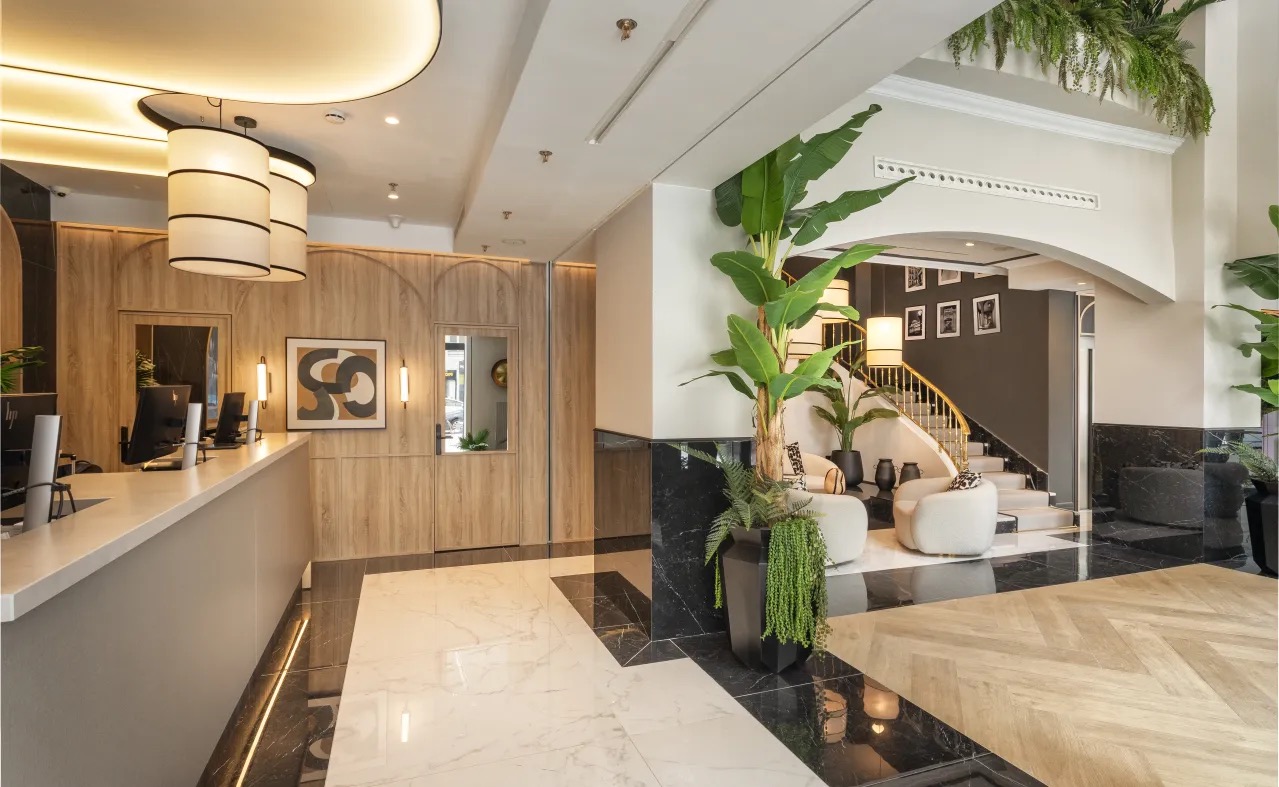 A hotel lobby with a wood-panelled reception desk, marble floor, tall potted banana plants and a curved staircase in the background.