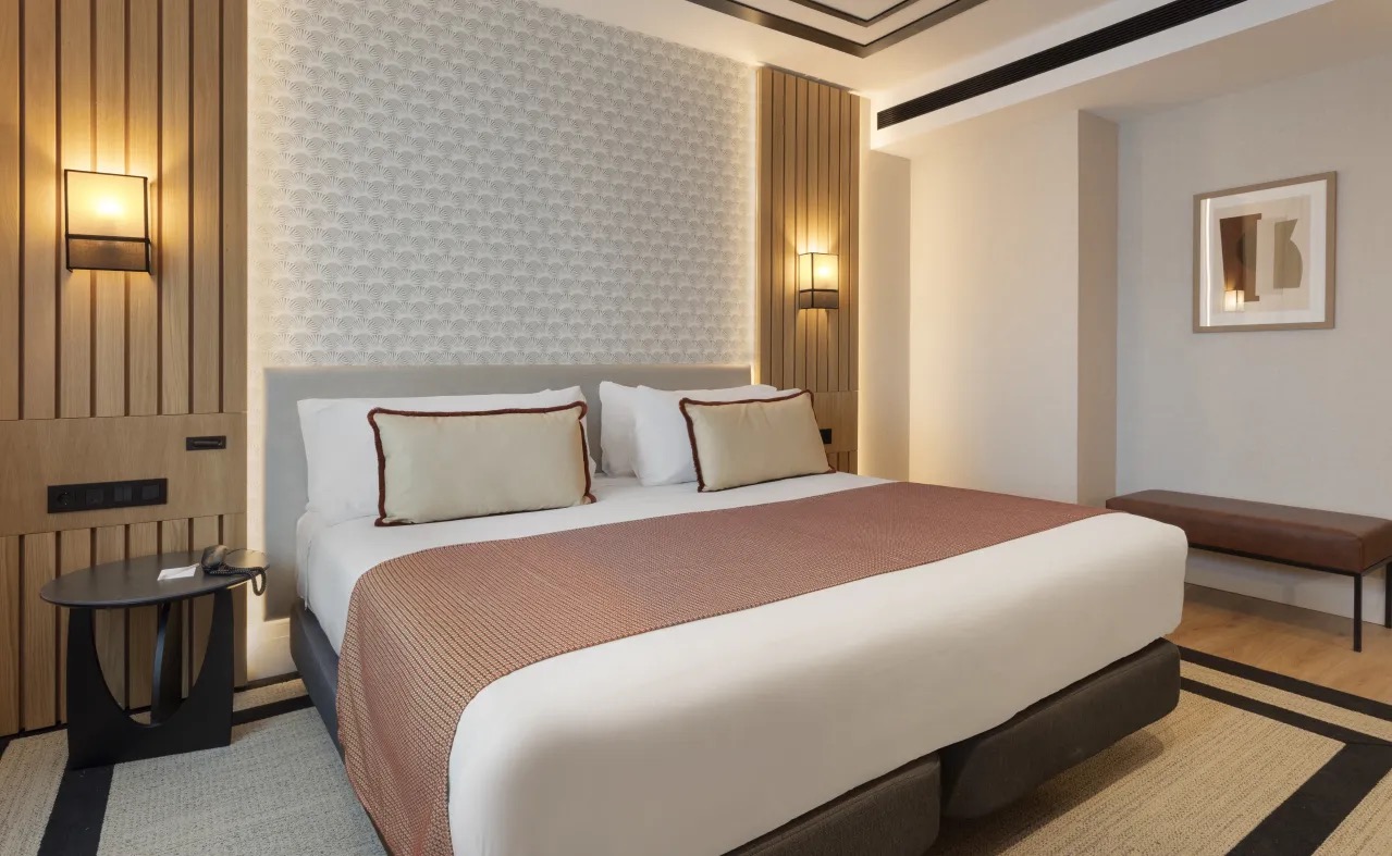 A hotel room with a patterned headboard and a small balcony with a bistro chair, overlooking the facade of a building across Gran Via.