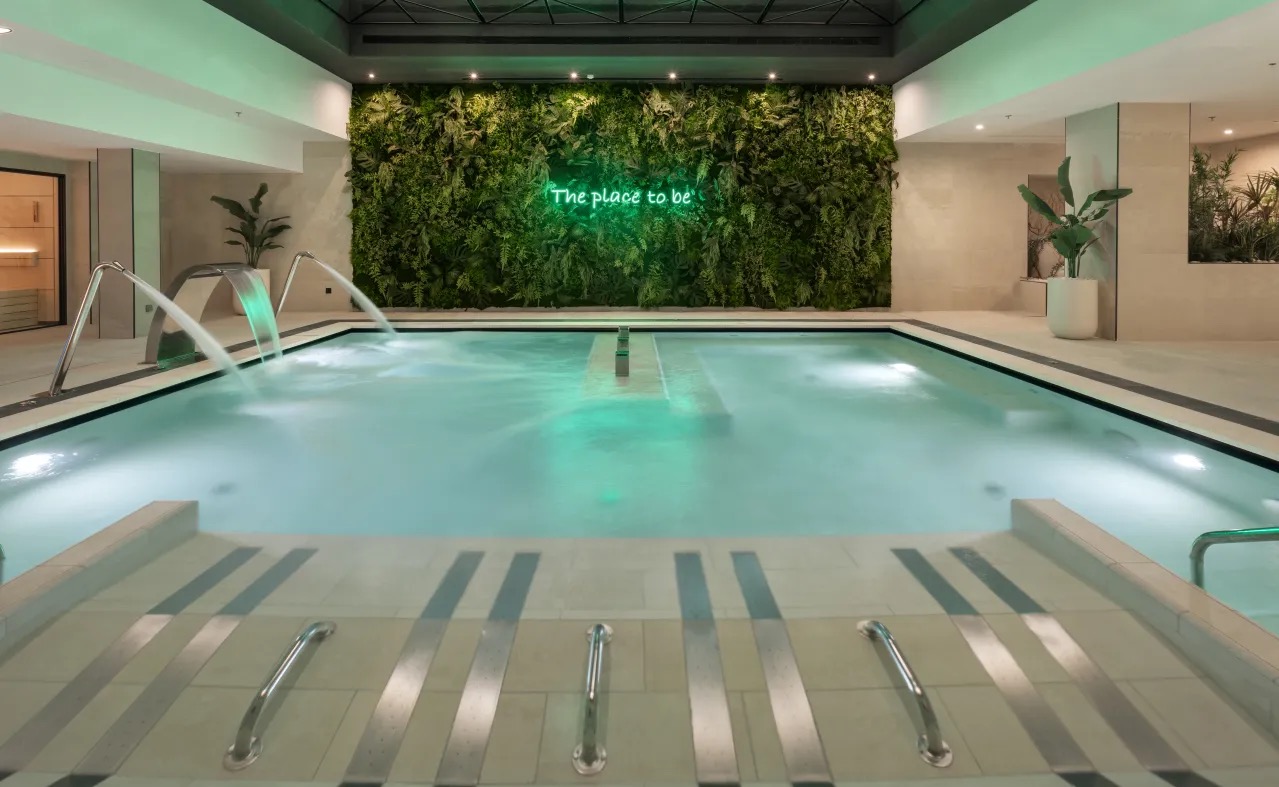 An indoor hydrotherapy pool lit in green, with jet fountains, a living plant wall and a neon sign reading The place to be.