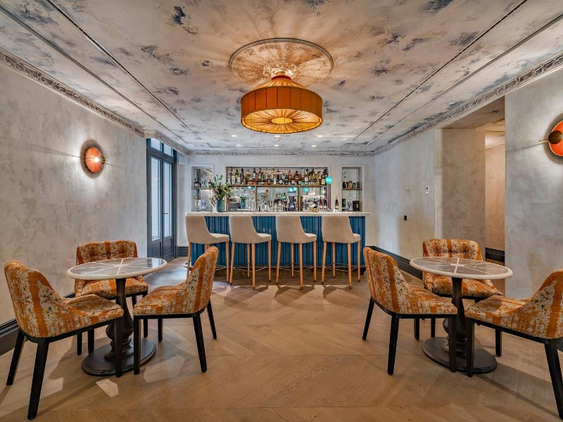 The pool-garden bar, with a painted fresco ceiling, a backlit shelf of bottles, and orange-patterned armchairs around small round tables.