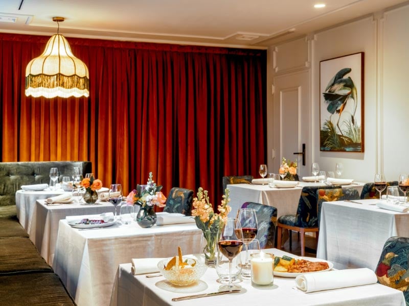 The El Patio de Atocha interior dining room, tables set with white linens and wine glasses beneath a fringed pendant lamp and a red velvet curtain.