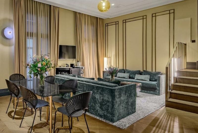 A suite's living and dining area, with a dark green velvet sofa, a glass dining table with six chairs, and a staircase to an upper level.