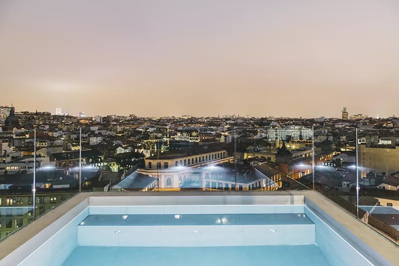 Rooftop pool empty at dusk, glass railing in the foreground and the illuminated Madrid skyline spread out behind it.