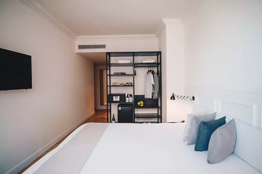 Family room with a wall-mounted TV, open wardrobe shelving holding folded towels and a minibar nook beside a white bed.