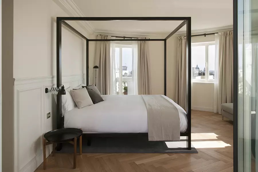 Suite bedroom with a black four-poster bed frame, herringbone wood floor and a balcony window looking over Madrid rooftops.