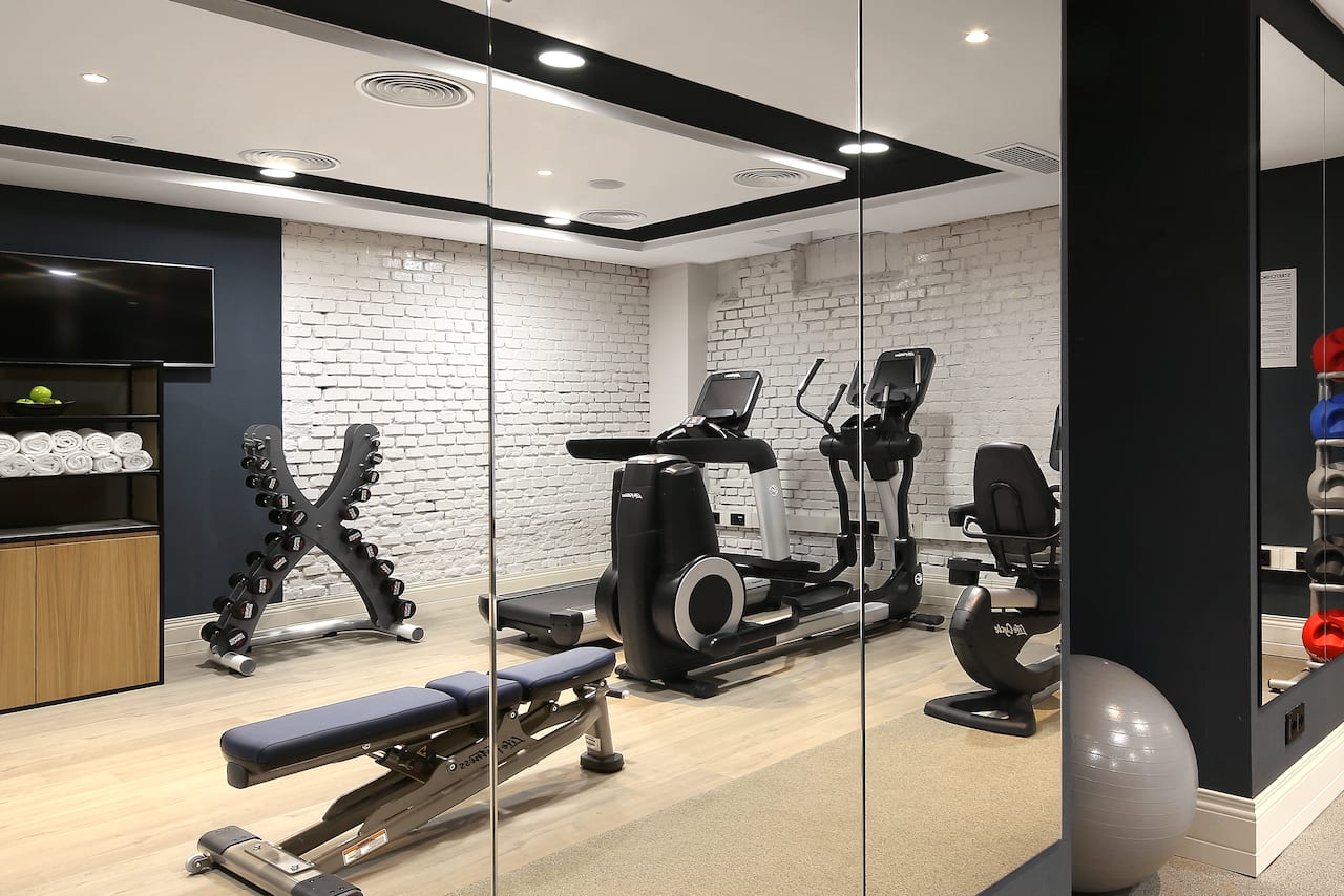 A small fitness room with a treadmill, elliptical machine, exercise bike, weight rack and exposed white brick wall.